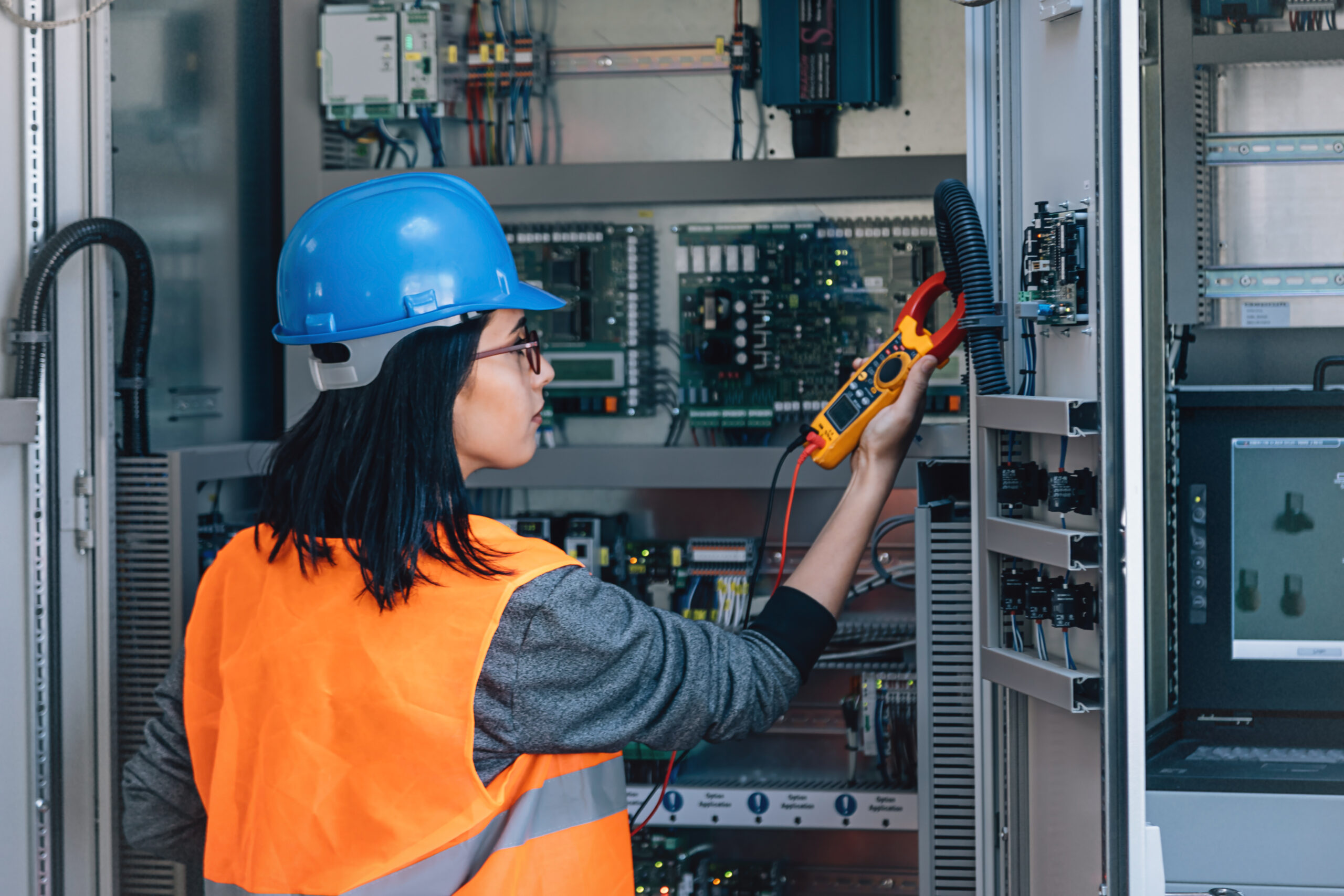 Young woman industrial service electrician engineer wearing protective vest and blue technician helmet testing and checking fridge electric voltage