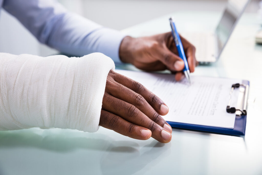Side View Of Injured Man With Bandage Hand Filling Insurance Claim Form On Clipboard for his personal injuries
