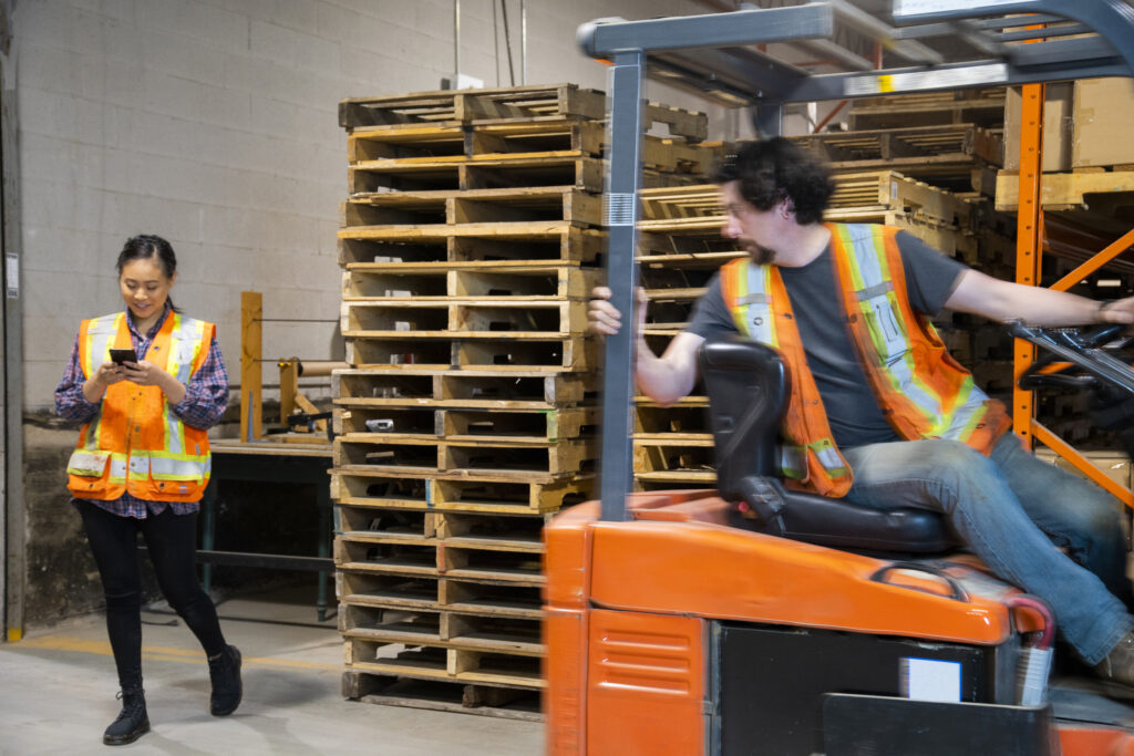 An industrial warehouse workplace safety topic. A female worker distracted by her mobile cell phone as a forklift approaches.  Accidents involving distracted employees and material handling vehicle
workers compensation