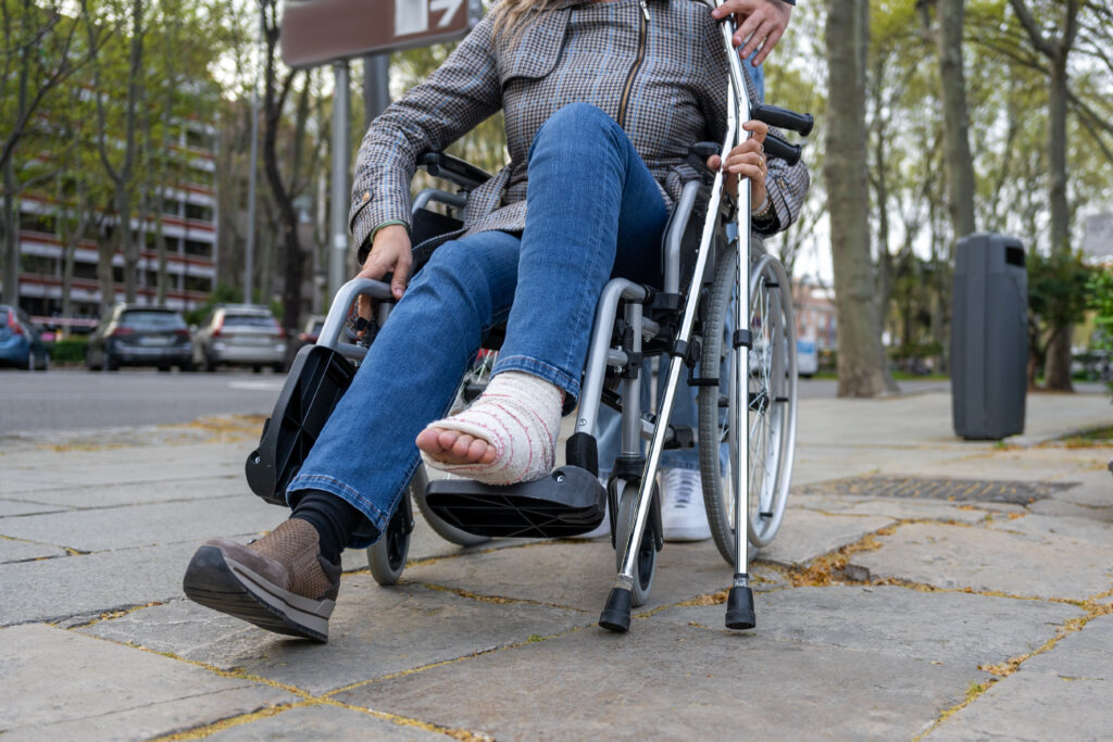 Low angle shot of an injured woman with a cast on her ankle, sitting in a wheelchair and carrying her crutches, in the middle of the street. work accident caused her injuries.