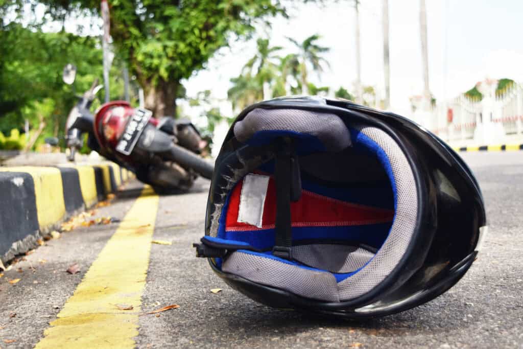 A motorcycle helmet sits on the street by the curb behind a crashed motorcycle further down the road. 